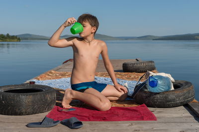 A boy slake his thirst with water from a mug, sitting on a wooden pier, against lake in summer day.
