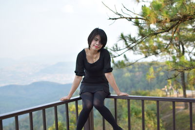 Portrait of young woman sitting railing against trees