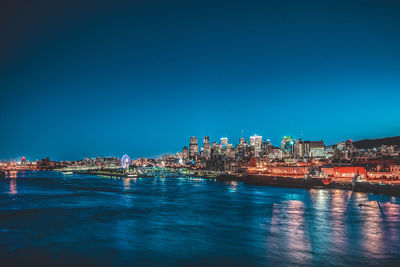 Illuminated buildings by river against blue sky at night