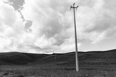 Low angle view of power lines on grassy hill against cloudy sky