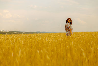 Portrait of young woman standing on wheat field against cloudy sky during sunset