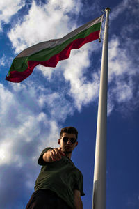 Low angle portrait of young man wearing sunglasses standing by flag against sky