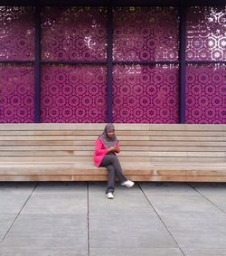 Woman wearing hijab using phone while sitting on bench against pink wall