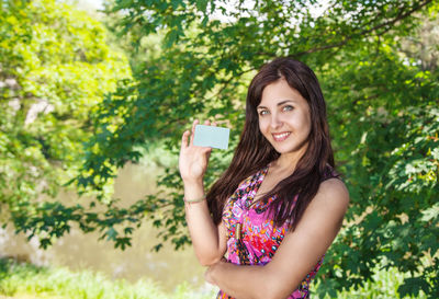 Portrait of smiling young woman holding card while standing against trees in park