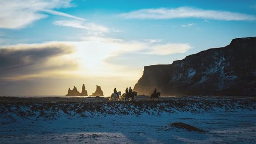 Silhouette people riding horses at beach against sky during winter