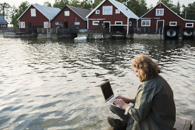 Man using laptop while sitting on jetty by lake against holiday villas