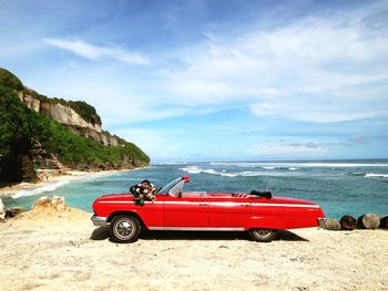 Red convertible at beach against cloudy sky