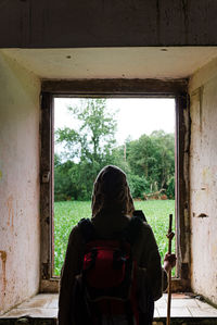 Back view of anonymous tired hiker with backpack leaning on stick while standing in doorway of aged building and resting after trekking in spanish countryside