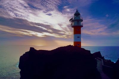 Lighthouse on cliff by sea against sky during sunset