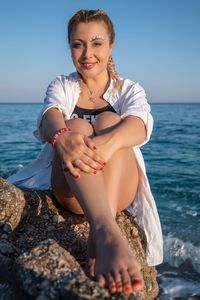 Young woman sitting on rock by sea against sky