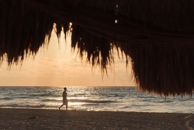 Man walking at beach in front of thatched roof during sunset