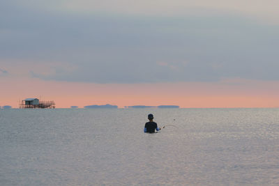 Rear view of man fishing in sea against sky during sunset