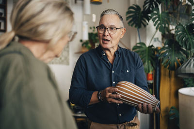 Senior male owner holding vase and talking with female customer doing shopping from antique store