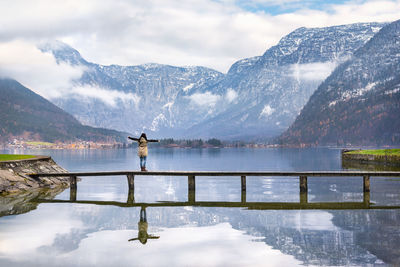 Rear view of woman standing with arms outstretched on bridge over lake hallstatter in hallstatt against mountains