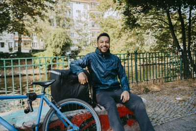 Portrait of happy male food delivery person sitting on bench by bicycle at park