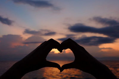 Cropped hands of woman forming heart shape against sea during sunset