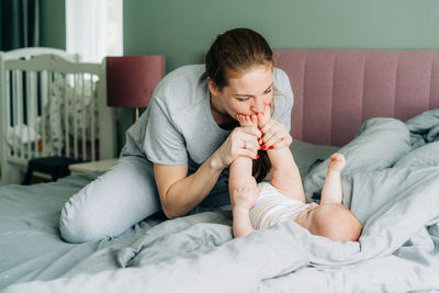 Young happy redhead mom is having fun, kissing the little feet of a newborn baby lying on the bed. 