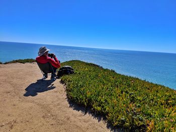 Rear view of man holding binoculars while sitting on chair at beach against sea and clear blue sky