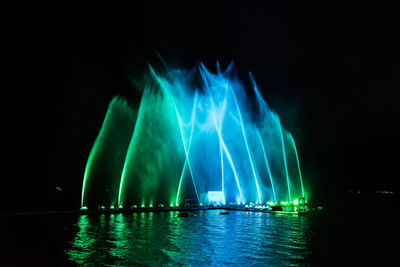 Illuminated water fountain by sea against clear sky at night