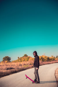 Side view of man with skateboard standing on road against clear blue sky