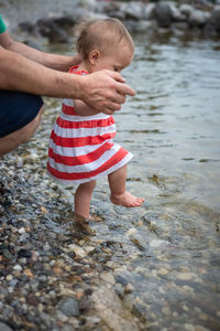 Close up view on baby in striped dress making steps in water being held by father hands