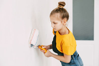 A girl in a denim overalls and a yellow t-shirt helps to paint the walls in an apartment white.