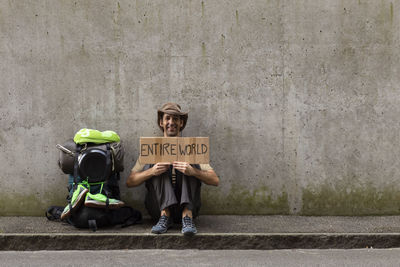 Portrait of man holding cardboard with entire world text while sitting against wall 