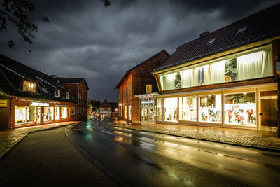 Wet street amidst illuminated stores against cloudy sky at night