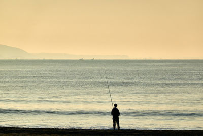 Silhouette man fishing in sea against sky during sunrise