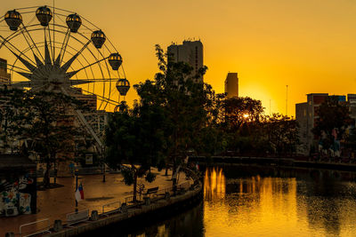 Ferris wheel by river against buildings in city at sunset