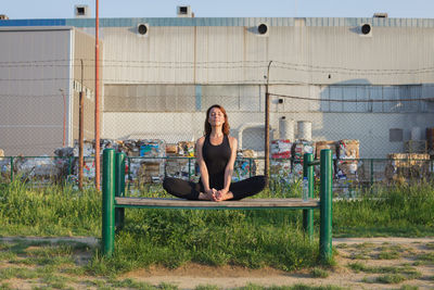 Woman practicing yoga on bench while sitting against factory