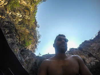 Portrait of shirtless man on rock against sky