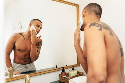 Side view of shirtless young man applying shaving cream on face while standing in bathroom