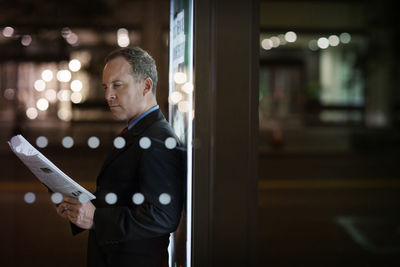 Serious businessman reading newspaper while standing against illuminated poster in city