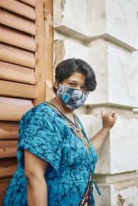 Portrait of a indian woman wearing face mask, standing near a heritage looking building exterior. 