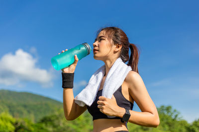 Young woman drinking water while standing against blue sky