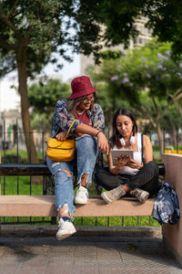 Full body of happy peruvian female friends in casual wear surfing tablet while sitting on bench on summer day in city