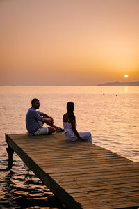 People sitting on shore by sea against sky during sunset