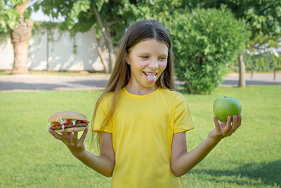 A teenage girl chooses between a burger and an apple outdoors in the park.