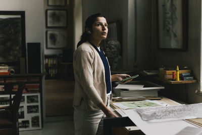 Contemplative female entrepreneur with blueprint and smart phone standing in home office