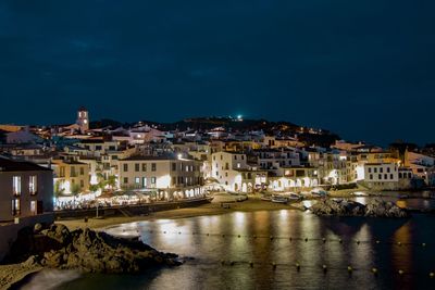 Illuminated buildings by sea against sky at night