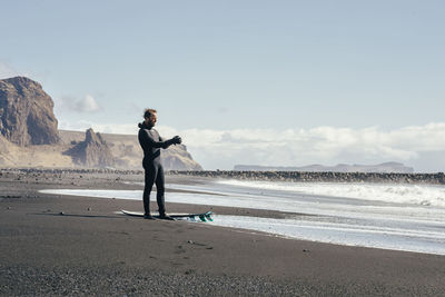 Man with surfboard standing at shore against sky during sunny day