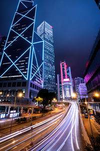 Light trails on road by illuminated buildings against sky at night