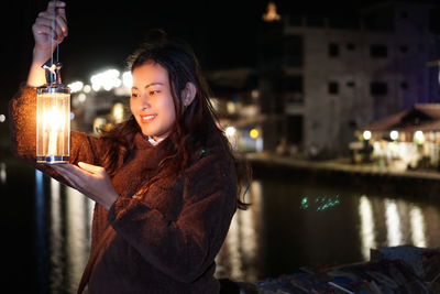 Portrait of young woman standing by river in city at night