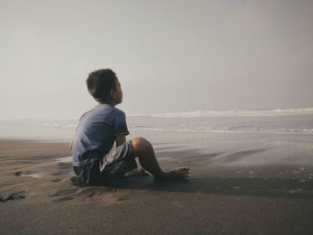 Rear view of man sitting on shore at beach against sky