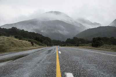 Empty road amidst grassy field against mountains during foggy weather