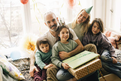 Portrait of happy birthday girl sitting with presents and family on alcove seat at home