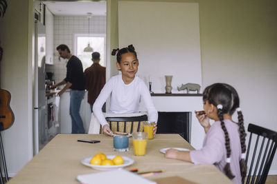 Smiling girl looking at sister while setting table for breakfast at home