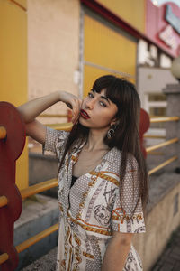 Young self esteem ethnic female with long dark hair in stylish outfit leaning on metal railing near modern building and looking at camera