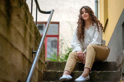 Portrait of smiling young woman sitting on staircase against wall